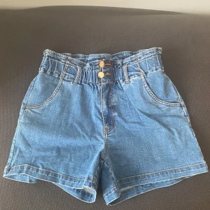 NÜMPH- Blå jeansshorts med hög midja - Snygga blå jeansshorts med hög midja och två knappar fram. De har en avslappnad passform och elastisk midja för extra komfort. Perfekta för sommardagar!