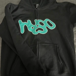 Svart hoodie med grön logga - Säljer en svart hoodie med en cool grön logga på framsidan och ett unikt mönster på baksidan. Tröjan har dragkedja och långa ärmar, perfekt för en avslappnad stil.