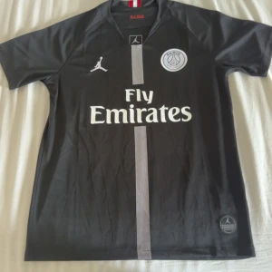 Svart PSG fotbollströja - Snygg svart PSG fotbollströja från Jordan med Fly Emirates-logga på framsidan. Tröjan har korta ärmar och ett stilrent mönster i mitten. Perfekt för fotbollsfans som vill ha en sportig look.