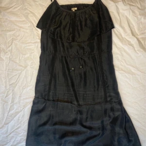 Svart klänning från Noa Noa - Elegant svart klänning från Noa Noa med volangdetaljer och knytband i midjan. Klänningen har en mjuk och glansig yta, perfekt för en stilren look. Axelbandslös design som ger en chic touch.