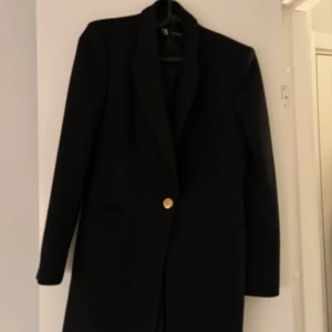 Svart kavaj med guldknapp - Elegant svart kavaj med en stilren design och en iögonfallande guldknapp framtill. Kavajen har långa ärmar och en klassisk krage, perfekt för en sofistikerad look.