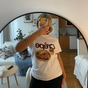 Vit t-shirt med crêpe-tryck - Säljer en vit t-shirt med ett stort tryck av en crêpe och texten 'Crêpe' i rött. Perfekt för dig som älskar mat och vill ha en rolig och unik stil. T-shirten är kortärmad och gjord i ett mjukt material.