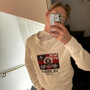 Vit sweatshirt från Morris - Säljer en stilren vit sweatshirt från Morris med ett stort tryck på framsidan som inkluderar flaggor och text. Tröjan har långa ärmar och en rund halsringning. Perfekt för en avslappnad stil.