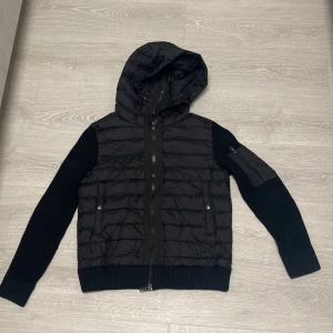 Moncler cardigan - Snygg svart pufferjacka från Moncler med stickade ärmar och huva. Jackan har dragkedja framtill och en praktisk ficka på ärmen. Perfekt för kyliga dagar!