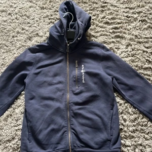 Blå hoodie från Sail Racing - Säljer en blå hoodie från Sail Racing med dragkedja och huva. Jackan har en praktisk bröstficka. Den är tillverkad i ett mjukt material som ger en bekväm känsla. Den är i bra skicka förutom att det är lite färg på luvan.