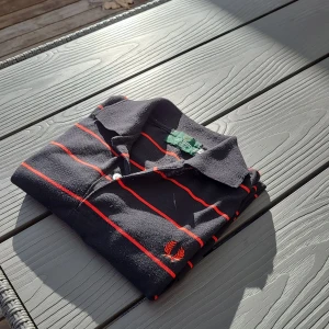 Grå och röd randig pikétröja från Fred Perry - Säljer en klassisk svart pikétröja från Fred Perry med röda ränder. Tröjan är tillverkad i 100% bomull och har korta ärmar samt en broderad logga på bröstet. Perfekt för en stilren look nu inför sommarn!