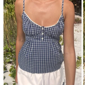 Blårutig linnetopp med knappar - Snygg blårutig linnetopp med smala axelband och knappar framtill. Toppen har en söt spetsdetalj längs halsringningen och en figurnära passform. Perfekt för varma sommardagar!