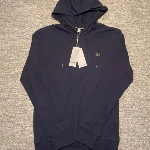 Mörkblå hoodie från Lacoste - Säljer en stilren mörkblå hoodie från Lacoste med den klassiska krokodilloggan på bröstet. Tröjan är tillverkad i mjuk bomull och har en justerbar huva med dragsko. Perfekt för en avslappnad look.