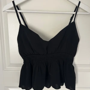 Svart topp - Säljer en snygg svart ärmlös topp med smala axelband och volangdetalj i nederkant. Toppen har en smockad midja som ger en fin passform. Perfekt för varma dagar eller som en del av en lager-på-lager-look.