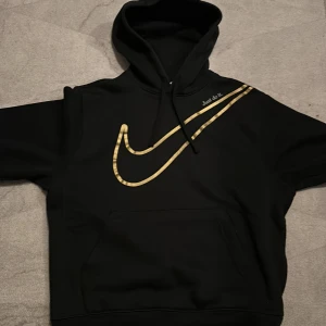 Svart hoodie från Nike - Säljer en svart hoodie från Nike med en stor guldfärgad logga på framsidan. Hoodien har en klassisk design med huva och långa ärmar. Storlek medium