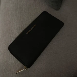 Svart plånbok från Michael Kors - Elegant svart plånbok från Michael Kors med dragkedja och guldiga detaljer. Perfekt för att hålla ordning på kort och kontanter. Stilren design som passar till alla tillfällen.