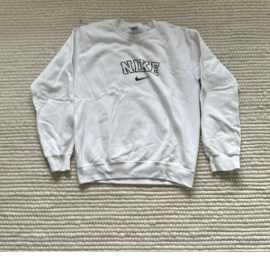 Vit sweatshirt från Nike - Säljer en klassisk vit sweatshirt från Nike med svart logotyp på bröstet. Tröjan har långa ärmar och ribbade muddar vid ärmslut och nederkant. Perfekt för en avslappnad stil.