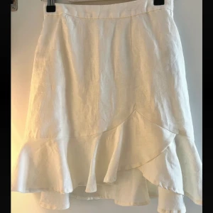 Linnekjol med volang - Säljer en stilren vit linnekjol med volangdetaljer. Kjolen har en omlott-look framtill och är perfekt för sommaren.