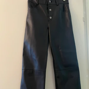 Svarta byxor från Zapa - Snygga svarta byxor från Zapa i ett skinnliknande material. Byxorna har hög midja och raka ben med knappar framtill. Perfekta för en stilren look.