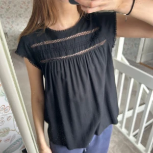 Svart blus med spetsdetaljer - Säljer en elegant svart blus med spetsdetaljer och korta ärmar. Blusen har en lös passform och en knapp i nacken för en stilren look. Perfekt för både vardag och festliga tillfällen. 