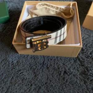  skärp med guldfärgat spänne - Säljer ett snyggt Burberry bälte i skinn med klassiskt rutig design i beige, svart och rött. Spännet är i guldfärgad metall med Burberrys ikoniska TB-logga. Bältet har en bred form och lyfter din outfit med lyxig känsla.