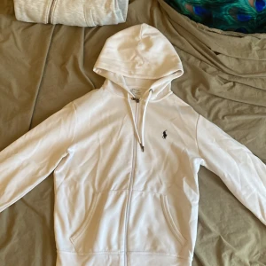 Ralph Lauren zip hoode - Vit hoodie från Polo Ralph Lauren med dragkedja och klassisk broderad logga på bröstet. Tröjan har huva med snörning och två fickor framtill. Materialet är en mix av polyester och bomull, vilket gör den både mjuk och bekväm.