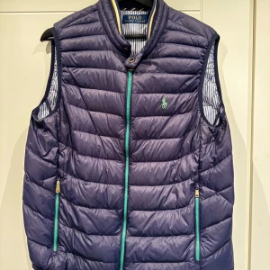 Mörkblå dunväst från Polo Ralph Lauren - Snygg mörkblå dunväst från Polo Ralph Lauren med grönt broderat logotyp på bröstet. Västen har quiltad design, två fickor med gröna dragkedjor och ståkrage. Insidan är fodrad med blå- och vitrandigt tyg. Perfekt för lager-på-lager-stil.