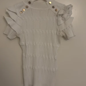 Vit stickad blus med volang från raglady - Supersöt vit stickad blus med coola guldknappar på axlarna och volangdetaljer vid ärmarna. Ribbad struktur och kortärmad modell som ger en trendig vibe. Perfekt för dig som vill sticka ut med en unik och stilren look.