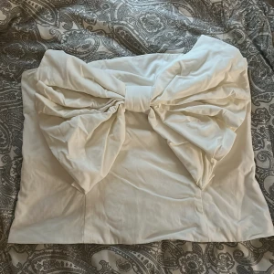 Vit axelbandslös tubtopp med rosett - Superfin vit axelbandslös tubtopp från Kappahl i storlek L. Toppen har en stor, iögonfallande rosett framtill som ger en unik och trendig look. Perfekt för dig som vill sticka ut och köra på något extra. Materialet känns slätt och har en lätt glans. Den är liten i storlek, jag har oftast S/M.