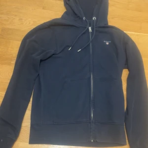 Marinblå hoodie från Gant - Snygg marinblå hoodie från Gant med dragkedja framtill och klassisk huva med snörning. Tröjan har två fickor på magen och diskret Gant-logga på bröstet. Perfekt för en chill och avslappnad stil.