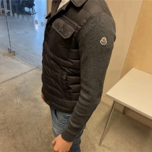 Svart cardigan från Moncler - Snygg svart dunväst från Moncler med quiltad front, två bröstfickor med lock och knappar samt klassisk Moncler-logga på ena ärmen. Västen har en krage och stängs med dragkedja. Ärmarna är i mörkgrå stickad ull för en schysst kontrast.