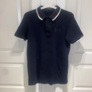 Svart EA7 polo t-shirt - Snygg svart polo t-shirt från EA7 Emporio Armani med vit kant på krage och ärmslut. Klassisk pikémodell med tre knappar framtill och diskret EA7-logga på bröstet. Perfekt för dig som gillar stilrena och sportiga plagg.