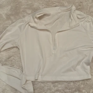 Vit croppad långärmad polotopp - Snygg vit croppad polotopp med lång ärm och halv dragkedja framtill. Mjuk och stretchig känsla, perfekt till jeans eller kjol. Enkel och clean design som funkar till många olika stilar.