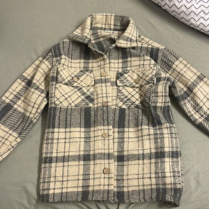 Rutig overshirt i beige och grå - Snygg och trendig overshirt i beige och grått rutigt mönster. Jackan har klassisk krage, två bröstfickor med knappar och långa ärmar. Perfekt att slänga på över en hoodie eller t-shirt för en chill look.