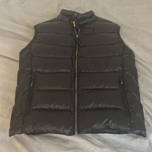 Parajumper Sheen Svart - Snygg svart dunväst från Parajumpers, modell Sheen. Västen har quiltad design, hög krage med tryckknapp och dragkedja framtill. Två dragkedjefickor på sidorna och klassisk PJS-logga bak i nacken. Perfekt för lager-på-lager under kyliga dagar. Skick 9/10.