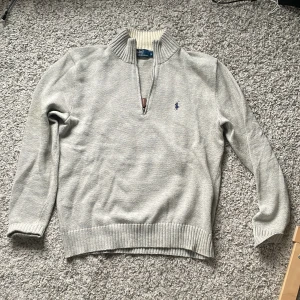 Grå stickad tröja Polo Ralph Lauren - Snygg grå stickad tröja från Polo Ralph Lauren i storlek S. Tröjan har half zip, ribbad krage och muddar samt den klassiska blå broderade loggan på bröstet. Tillverkad i 100% bomull, perfekt för kyliga dagar och enkel att matcha med jeans eller chinos.