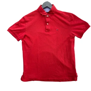 Röd polo t-shirt Tommy Hilfiger M - Snygg röd polo t-shirt från Tommy Hilfiger i klassisk passform. T-shirten har korta ärmar, krage och knappar framtill. Materialet är mjuk bomull och den har en liten Hilfiger-logga på bröstet. Perfekt för dig som gillar stilrena och ikoniska plagg. Priset är inte hugget i sten. Skriv om ni har några frågor! //Mvh Arvid!