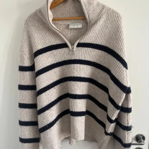 Randig stickad half zip-tröja Neo Noir - Beige stickad tröja från Neo Noir med svarta horisontella ränder. Tröjan har en hög krage med half zip-dragkedja och långa ärmar. Oversized och mysig passform, perfekt för kyliga dagar. Ribbstickad struktur ger en trendig och avslappnad look.