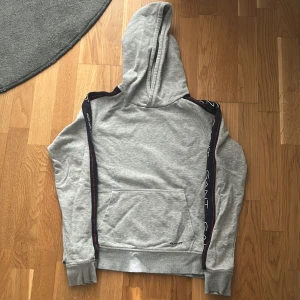 Grå hoodie från GANT med logoband - Grå hoodie från GANT med huva och känguruficka. Svarta logoband med GANT-tryck längs ärmarna och diskret logga framtill. Mjuk bomullskänsla och ribbade muddar. Perfekt för chill dagar eller till skolan.