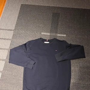 Mörkblå Tommy Hilfiger sweatshirt - Snygg mörkblå sweatshirt från Tommy Hilfiger med diskret logga på bröstet och klassisk rund hals. Tröjan har långa ärmar och ribbade muddar vid ärmslut och nederkant. Perfekt för dig som gillar stilren och enkel design.