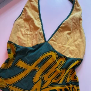 Gul och grön halterneck linne💕 - Retroinspirerad halterneck linne i gult och grönt med djup v-ringning och tryckt text i sportig stil. Toppen har breda axelband som knyts i nacken och är perfekt för dig som vill sticka ut med en färgglad och unik look. Skriv vid frågor❤️🥰