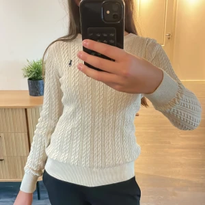 Beige kabelstickad tröja Polo Ralph Lauren - Kabelstickad vit tröja från Polo Ralph Lauren med klassisk marinblå logga på bröstet. Tröjan har rund hals, ribbade muddar och är långärmad.