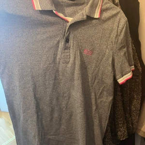(helt ny) Grå Boss pikétröja med rosa detaljer  - Snygg grå pikétröja från Hugo Boss med regular fit. Tröjan har korta ärmar och krage med rosa och vita ränder, samt rosa Boss-logga på bröstet. Materialet är mjuk bomull som känns skönt mot huden. Perfekt för dig som gillar stilrena och sportiga plagg.