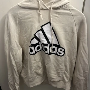 Vit hoodie från Adidas med logga - Snygg vit hoodie från Adidas med stor svartvit logga på bröstet. Tröjan har huva med snörning och långa ärmar. Tillverkad i mjuk bomullsmix som är skön att ha på sig. Perfekt för en sportig och avslappnad stil.