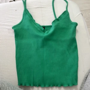 Grön ribbad linnetopp med v-ringning - Snygg grön ribbad linnetopp med v-ringad hals och smala axelband. Toppen är croppad och har en vågig kant nedtill som ger en cool touch. Perfekt att matcha med jeans eller kjol för en chill look.