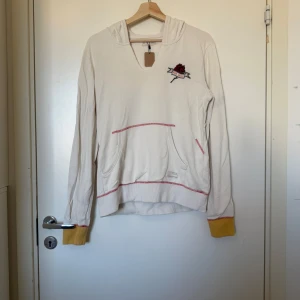 Vit hoodie med broderad ros och gula muddar - Cremevit/ ljus beige hoodie Odd Molly med broderad ros och text på bröstet. Har röda sömmar som kontrast, gula muddar vid ärmslut och praktisk känguruficka framtill. Mjuk bomullskänsla och huva för extra chill vibe. Perfekt för dig som gillar unika detaljer. Storlek 2 som enligt deras storleksguide motsvarar en storlek M. Fint skick, men se bild 3 för anmärkning. 