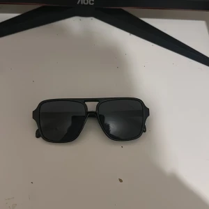 Svarta solglasögon med fyrkantig form - Snygga svarta solglasögon med fyrkantiga linser och bred båge. De har en modern och stilren design med helsvart plastmaterial och mörka glas. Perfekta för att ge en cool vibe till din outfit.