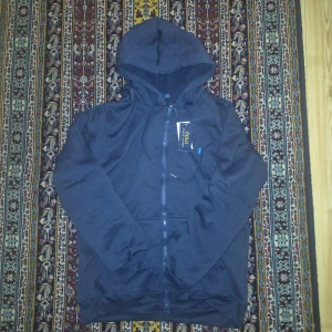 Mörkblå hoodie från Polo Ralph Lauren - Mörkblå hoodie från Polo Ralph Lauren med dragkedja framtill och klassisk huva med snörning. Tröjan har långa ärmar, ribbade muddar och är tillverkad i mjuk bomull. Perfekt för en chill och avslappnad stil.