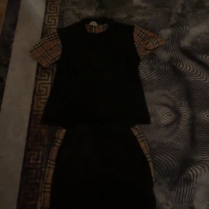 Burberry kort klänning med rutiga detaljer - Snygg kort klänning från Burberry med klassiska beige och svarta rutiga detaljer på ärmar och sidor. Klänningen är svart med korta ärmar och har en loose passform. Perfekt för dig som vill sticka ut med en ikonisk look.