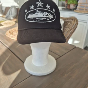 Svart truckerkeps Cortez - Svart truckerkeps från Cortez med vit brodyr framtill och på sidorna, prydd med stjärnor och motiv. Kepsen har mesh på sidorna och bak, samt justerbar snapback. Märkt 'Cortez Rules the World' på insidan. Perfekt för dig som gillar streetstyle.