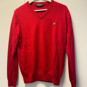 Röd v-ringad tröja Lyle & Scott - Snygg röd v-ringad tröja från Lyle & Scott med deras klassiska gula fågel-logga på bröstet. Tröjan är långärmad och har ribbade muddar vid ärmslut och nederkant. Perfekt för dig som gillar stilrena och enkla plagg med en touch av färg.