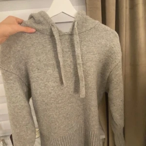 Grå stickad hoodie med snörning - Mysig grå stickad hoodie med huva och snörning. Tröjan har långa ärmar och bred ribbad kant nedtill för en snygg och avslappnad look. Perfekt för kyliga dagar när du vill vara både bekväm och stilren. 