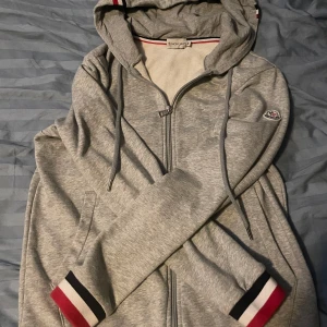 Grå Moncler hoodie med dragkedja - Snygg grå hoodie från Moncler med dragkedja och huva. Tröjan har ribbade muddar med röd, vit och svart detalj samt Moncler-logga på ärmen. Perfekt för dig som gillar stilrena och sportiga plagg med premiumkänsla.