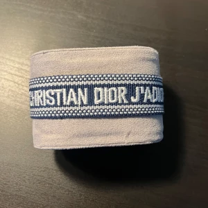 Blå Christian Dior J'Adior armband - Snyggt blått armband från Christian Dior med texten 'J'Adior' broderad i vitt. Armbandet är gjort i tyg och har coola tofsar i ändarna samt justerbar knytning. Perfekt för att ge din outfit en lyxig och trendig touch.