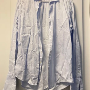 Ljusblå klassisk bomullsskjorta - Snygg ljusblå skjorta i bomull med klassisk krage och långa ärmar. Skjortan har vita knappar framtill och vid manschetterna. Passformen är normal och tyget är mjukt och bekvämt. Perfekt för dig som gillar stilrena och enkla plagg.